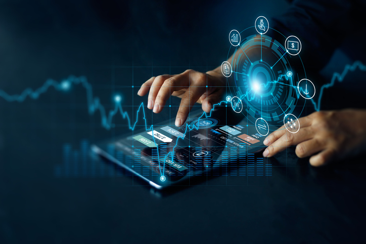 Hands type on a tablet with blue holographic financial icons and chart visuals projecting above the device.