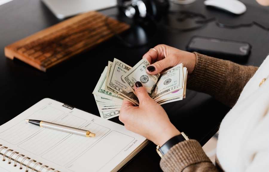 Person counting a stack of US dollars over an open planner on a dark desk.