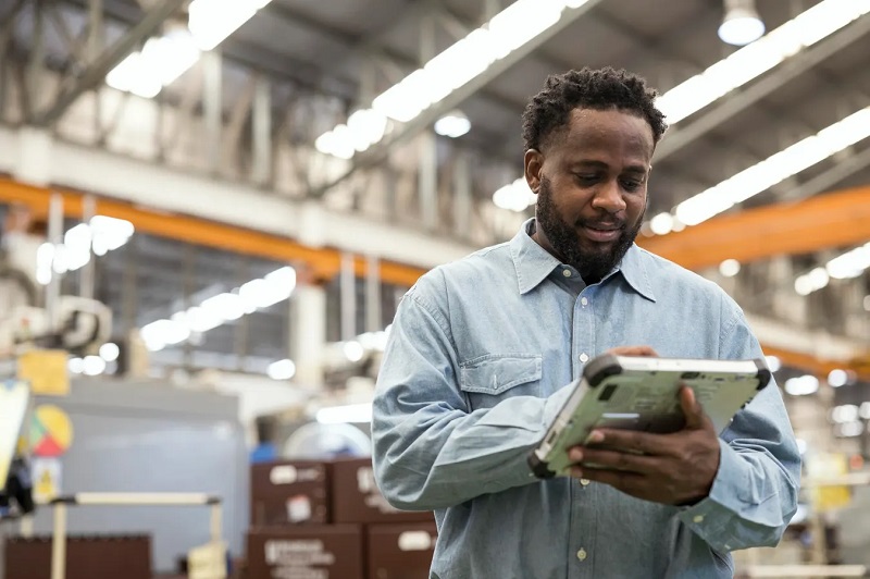 A man in a light blue work shirt uses a tablet in a busy factory warehouse.