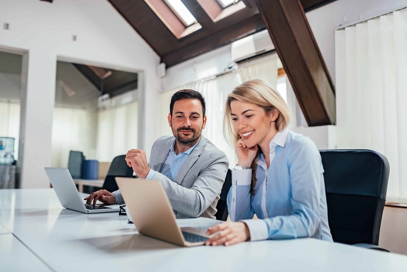 Two coworkers in business attire collaborate at a bright conference room table, looking at laptops and smiling.